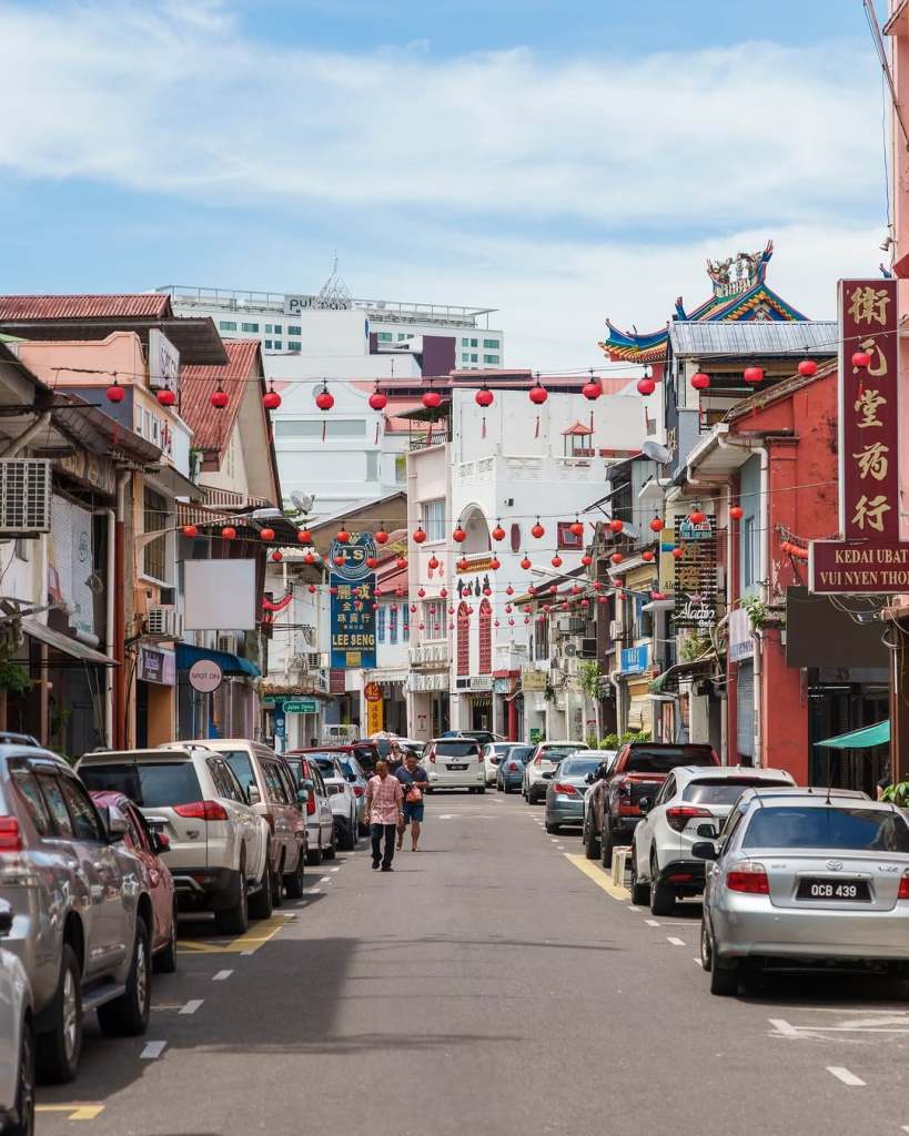 A bustling street scene in Kuching with colorful traditional lanterns overhead, featuring parked cars lining both sides of the road and pedestrians walking along the street.