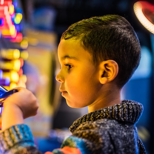 Young child in a striped sweater intently focuses on colorful lights and buttons, possibly playing an arcade game.