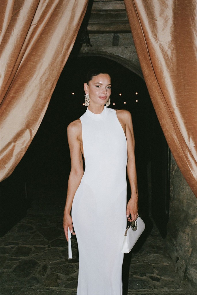 Bride in a minimalist white evening dress standing beneath draped curtains at night