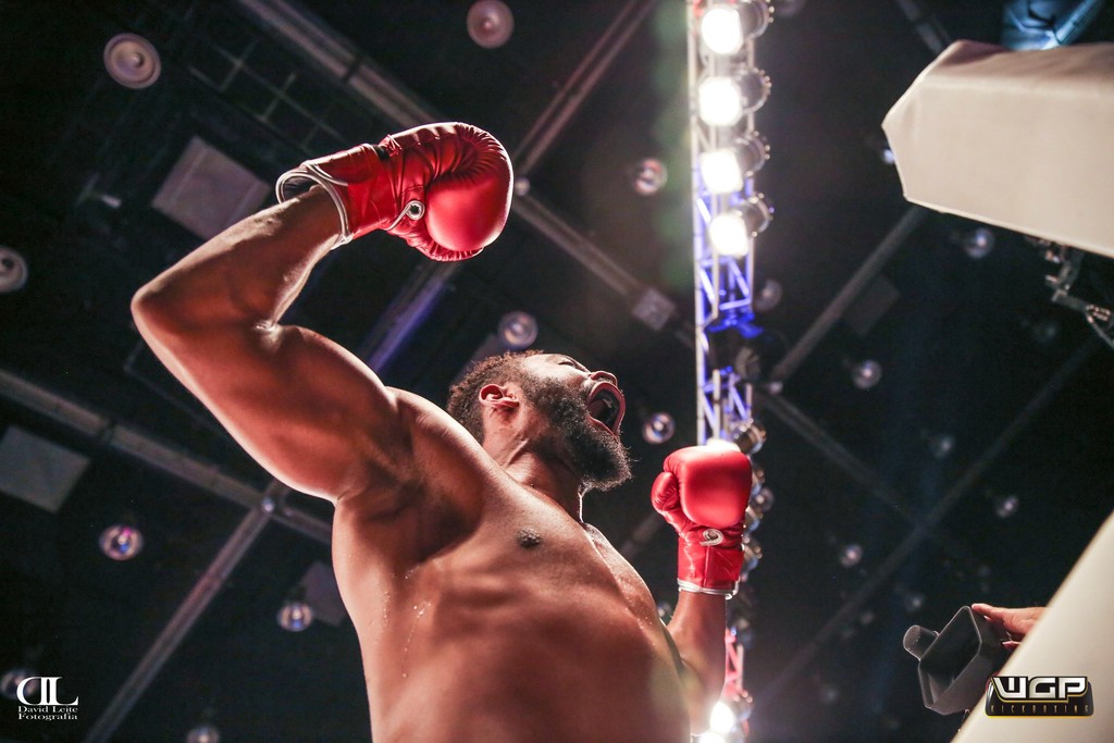 A boxer raises his gloves in victory, with bright lights and an arena setting in the background.