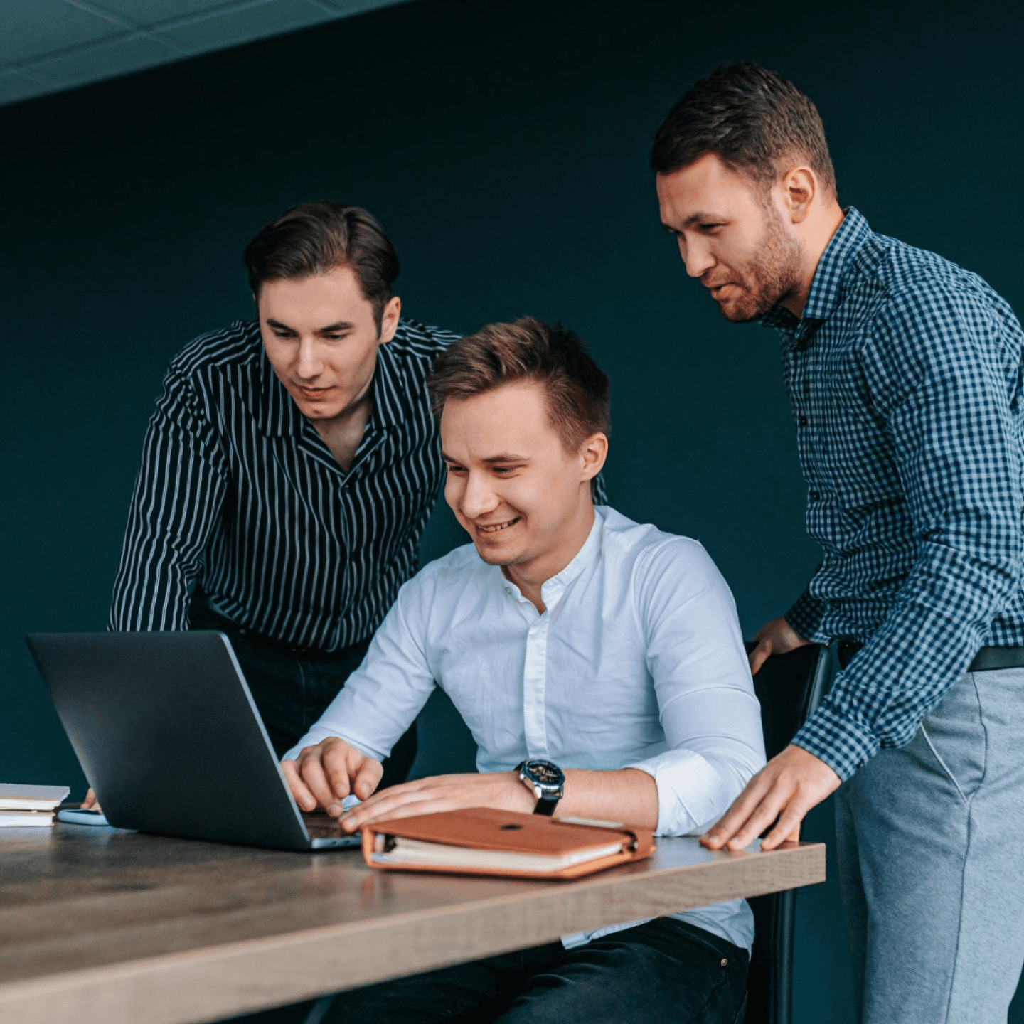 Three people gathered around a laptop, smiling and discussing in a modern office