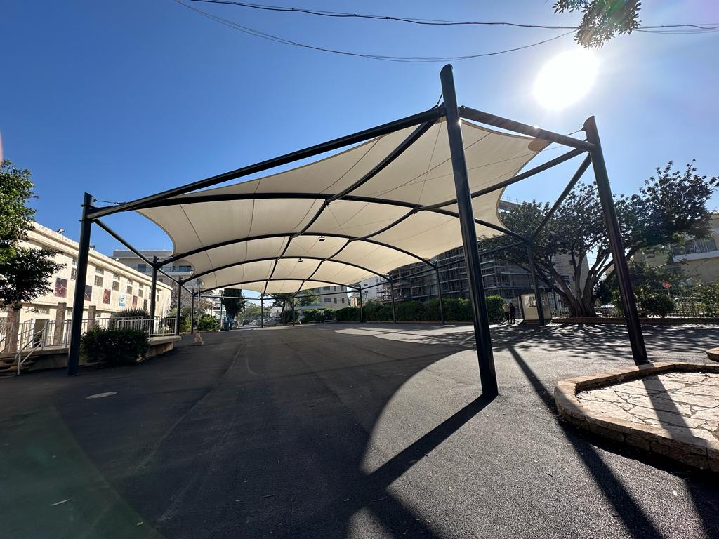 Large beige sunshade canopy on black metal frame over paved outdoor area under sunny sky.