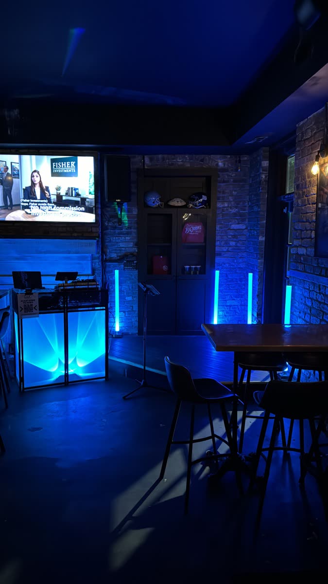 DJ booth with a laptop and controller set up in a brick-walled bar, lit with blue and teal LED uplighting