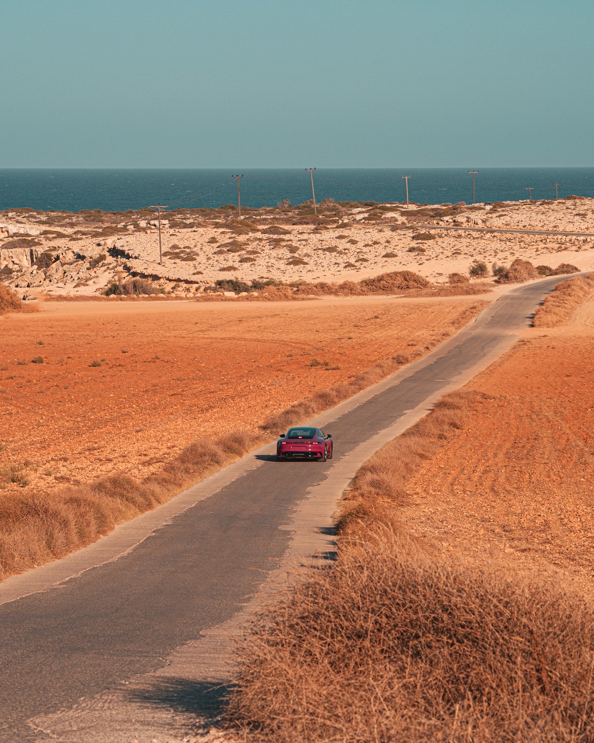 Ruby Star Porsche 911 Carrera T (992) seen from behind, small in frame, driving along a narrow road through arid orange scrubland toward the sea on the horizon, in Cyprus.