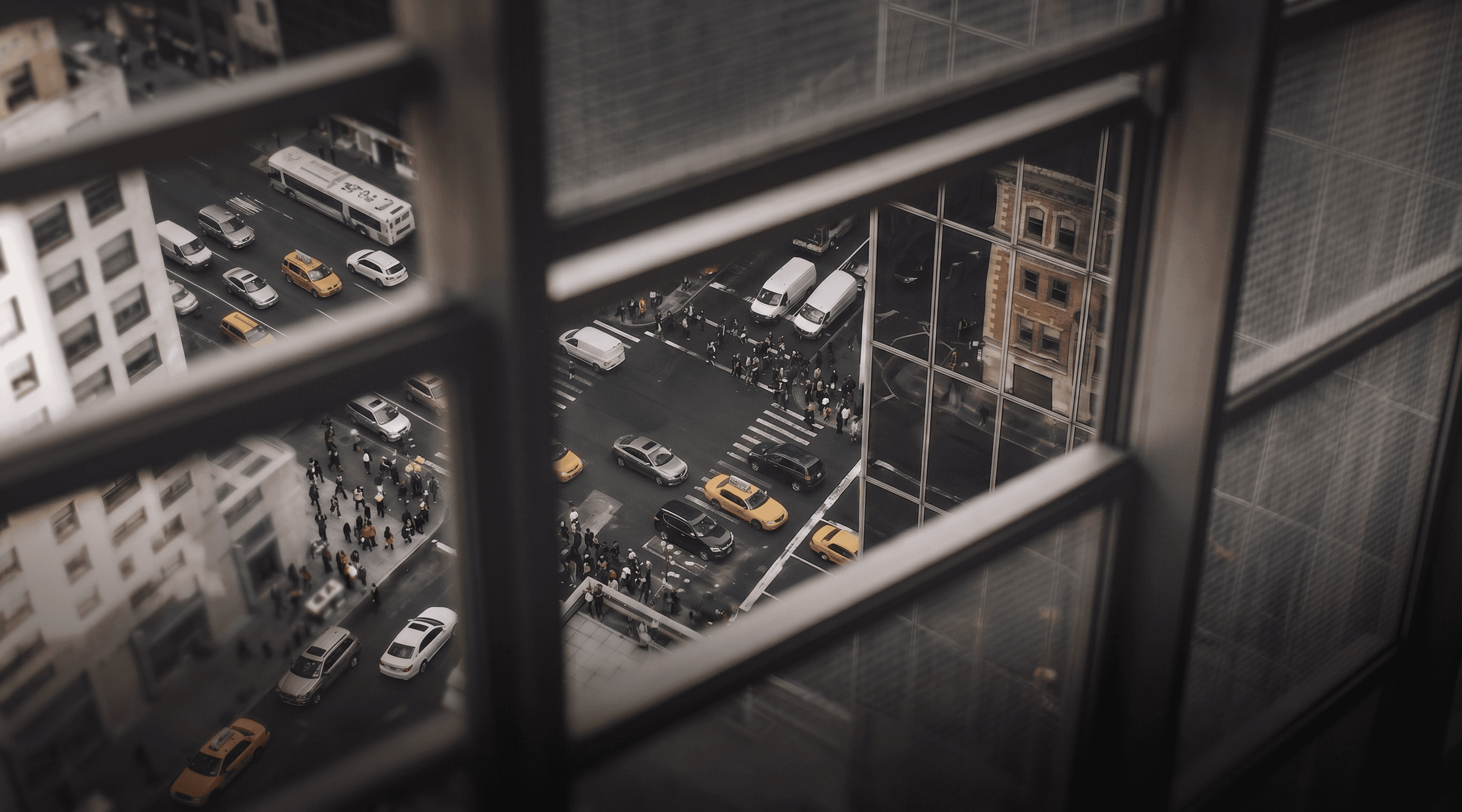 Aerial view of a busy city intersection with cars, yellow taxis, a bus, and pedestrians crossing the street, seen through a grid window frame.