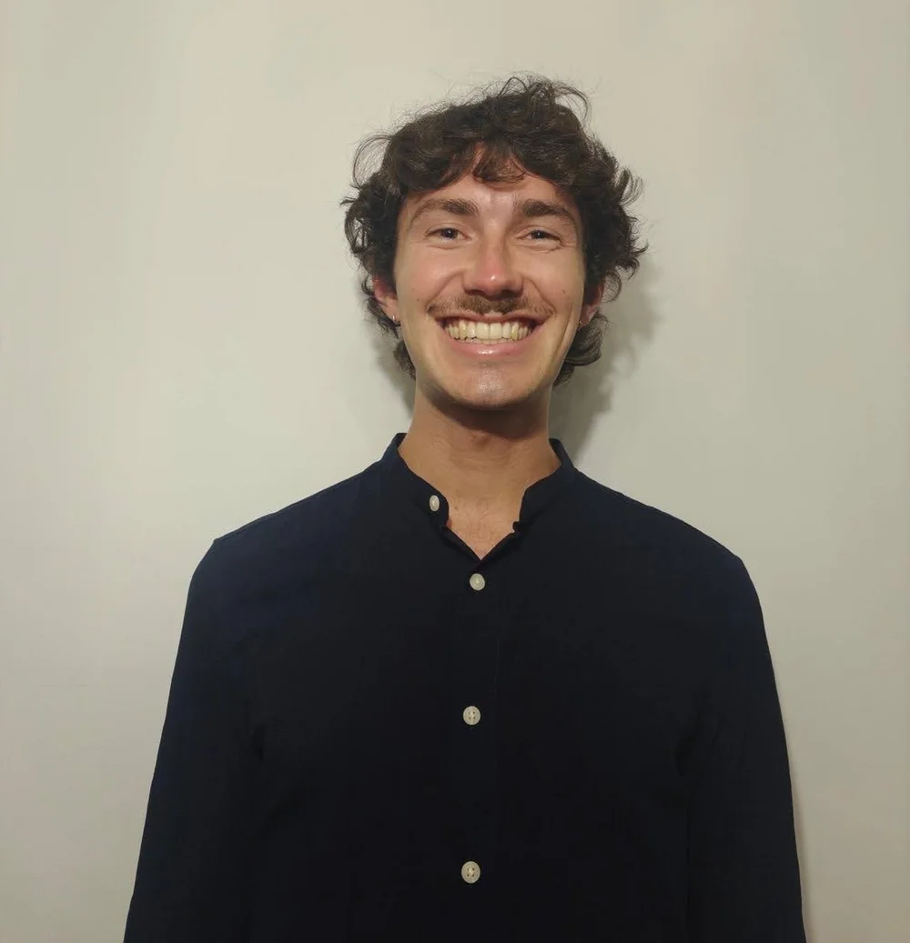 Uma pessoa com cabelo castanho cacheado e um sorriso largo está vestindo uma camisa preta de botões, diante de um fundo branco liso.