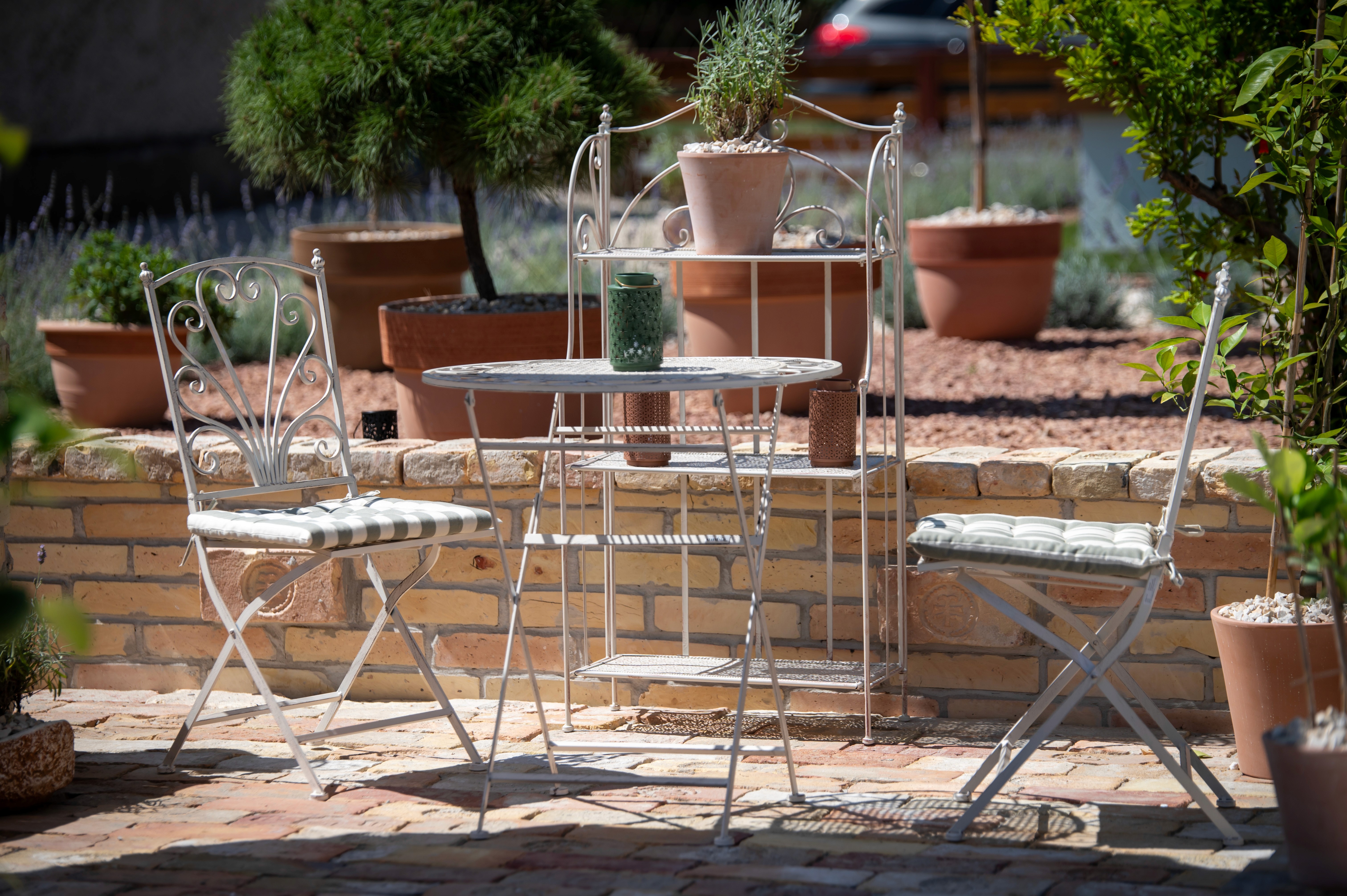 Hangulatos kültéri terasz két díszes fém székkel és egy kerek asztallal, kőburkolaton elhelyezve, buja cserepes növényekkel és téglafallal körülvéve, meghitt kerti hangulatot teremtve.