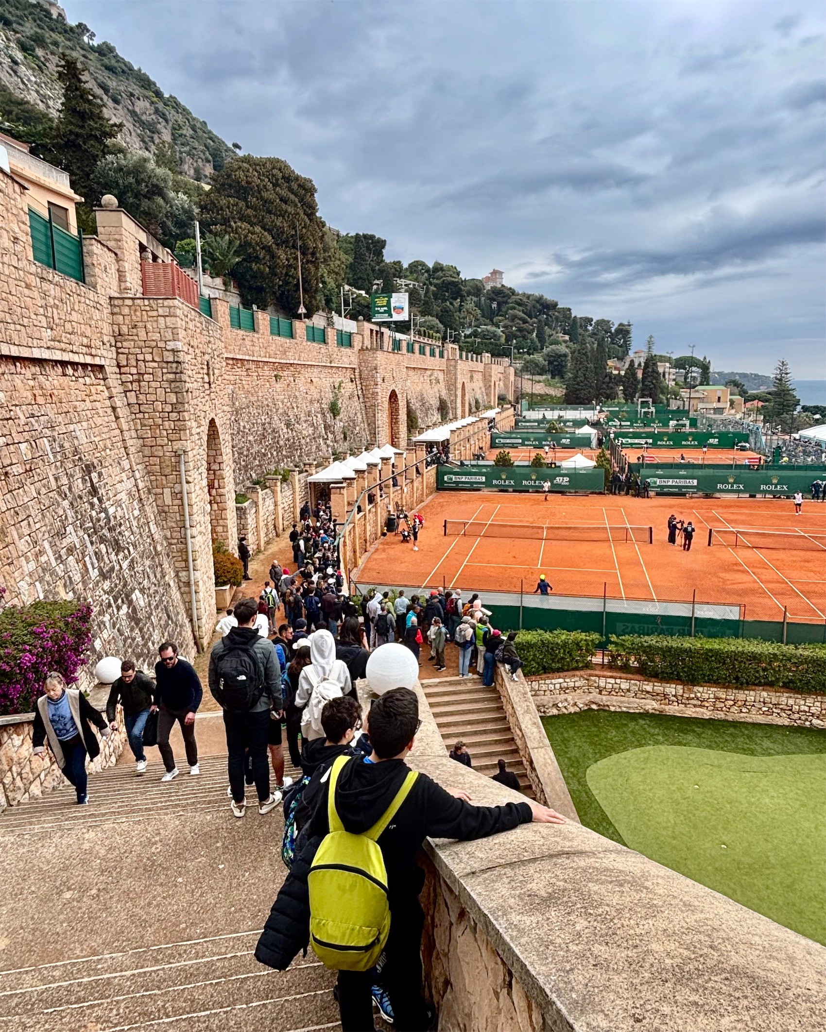 Red clay courts terraced below a stone wall at the Monte-Carlo Country Club, with the sea beyond