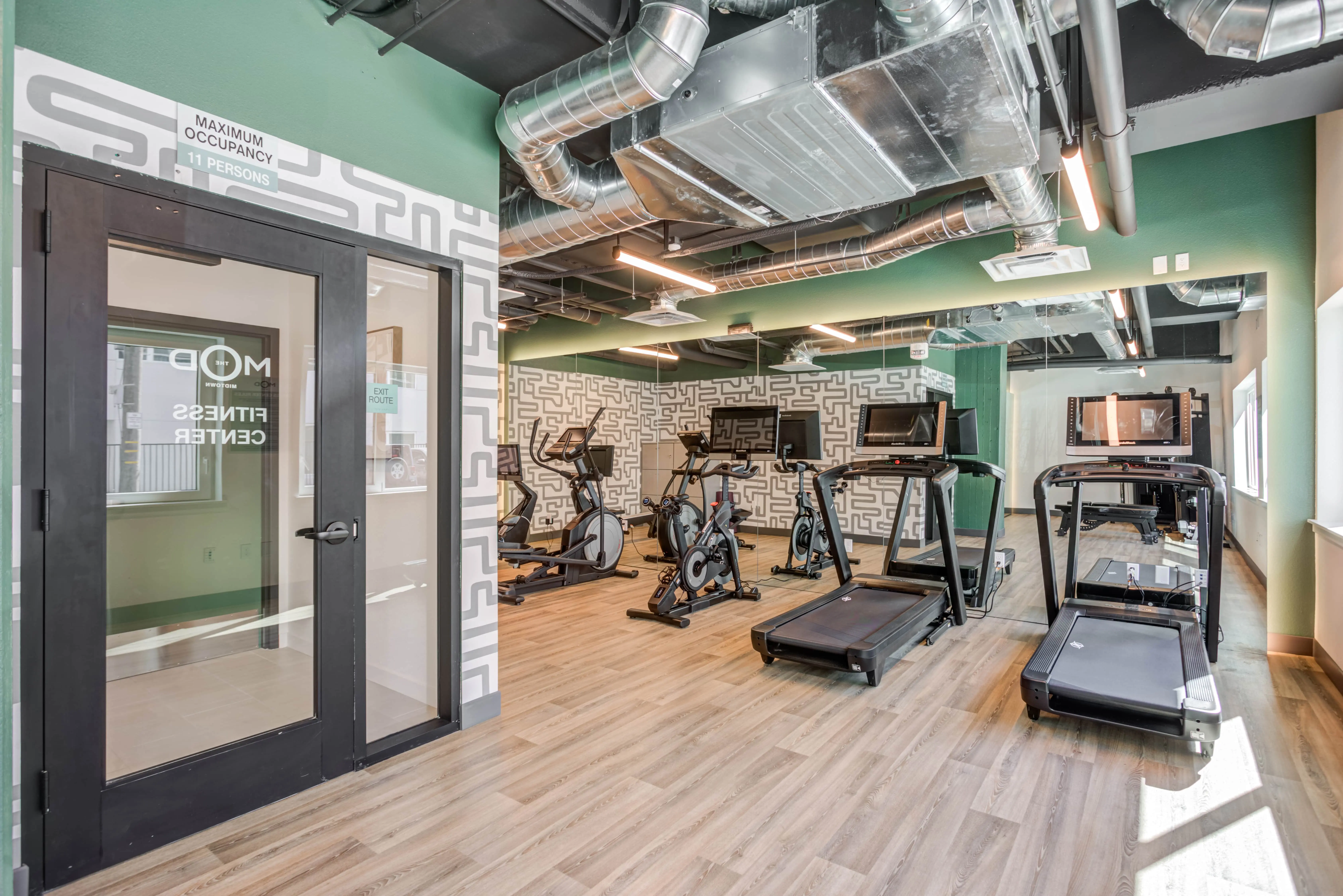 A modern fitness center in Midtown Sacramento with large mirrors and wooden flooring. The sun-drenched room contains treadmills, stationary bikes, and other cardio equipment. An adjoining room with glass doors has a sign that reads "Maximum Occupancy 15 Persons." Air ducts are visible on the ceiling.