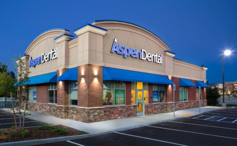 Exterior of an Aspen Dental office during early evening, with bright signage, blue awnings, and large promotional windows advertising free exams and X-rays. Highlights the welcoming and accessible design of Aspen Dental clinics.