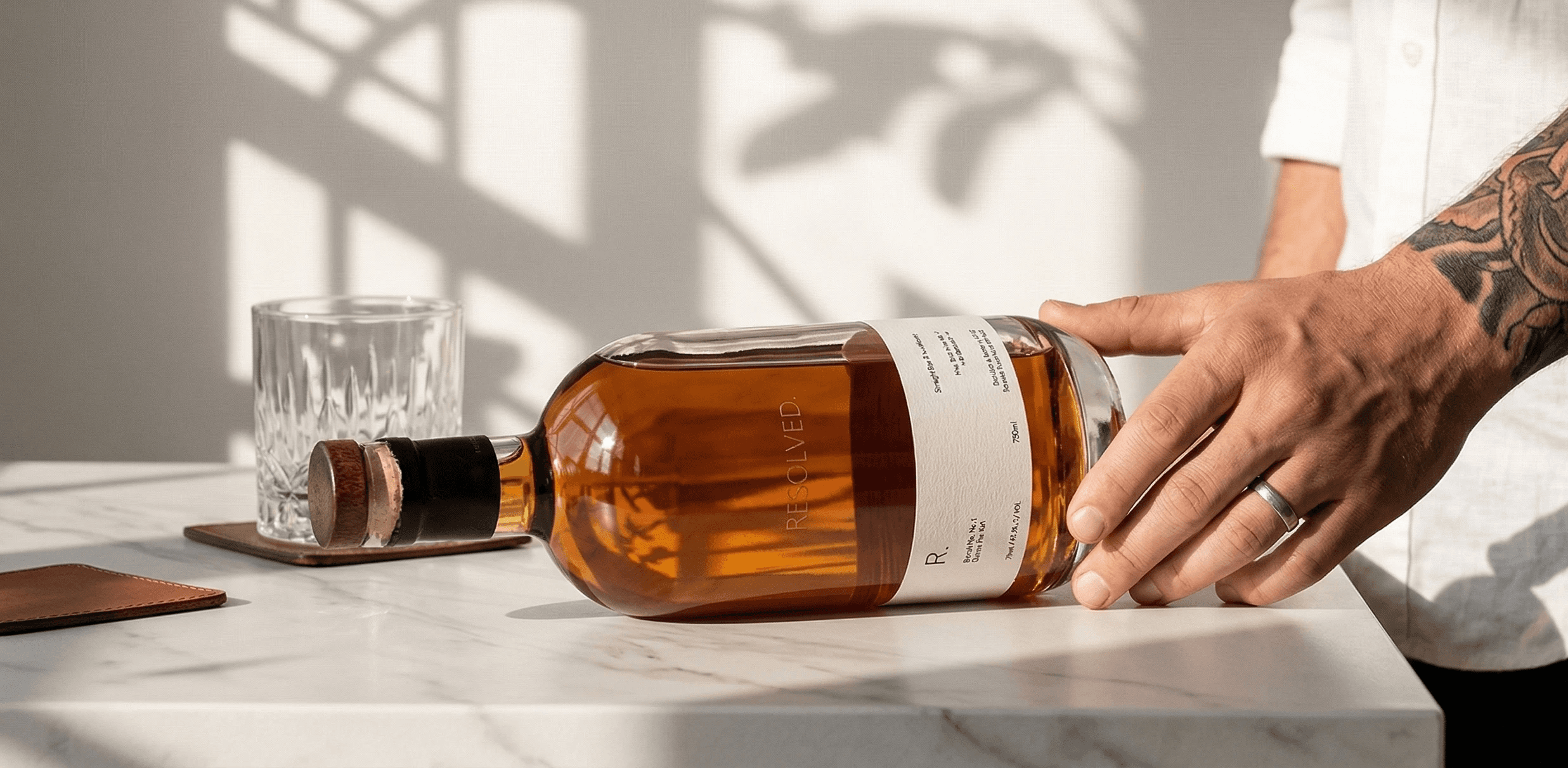 A hand with a tattoo gently places a bottle of REOLVED. whiskey on a marble countertop, accompanied by an empty glass ready for pouring, with soft, natural light casting shadows in the background.