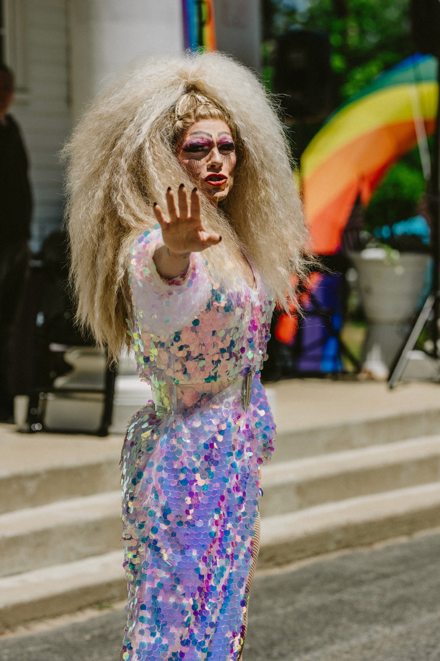 A drag performer in full look reaches toward the camera on the Halifax Town Hall steps, a rainbow flag draped in the background.