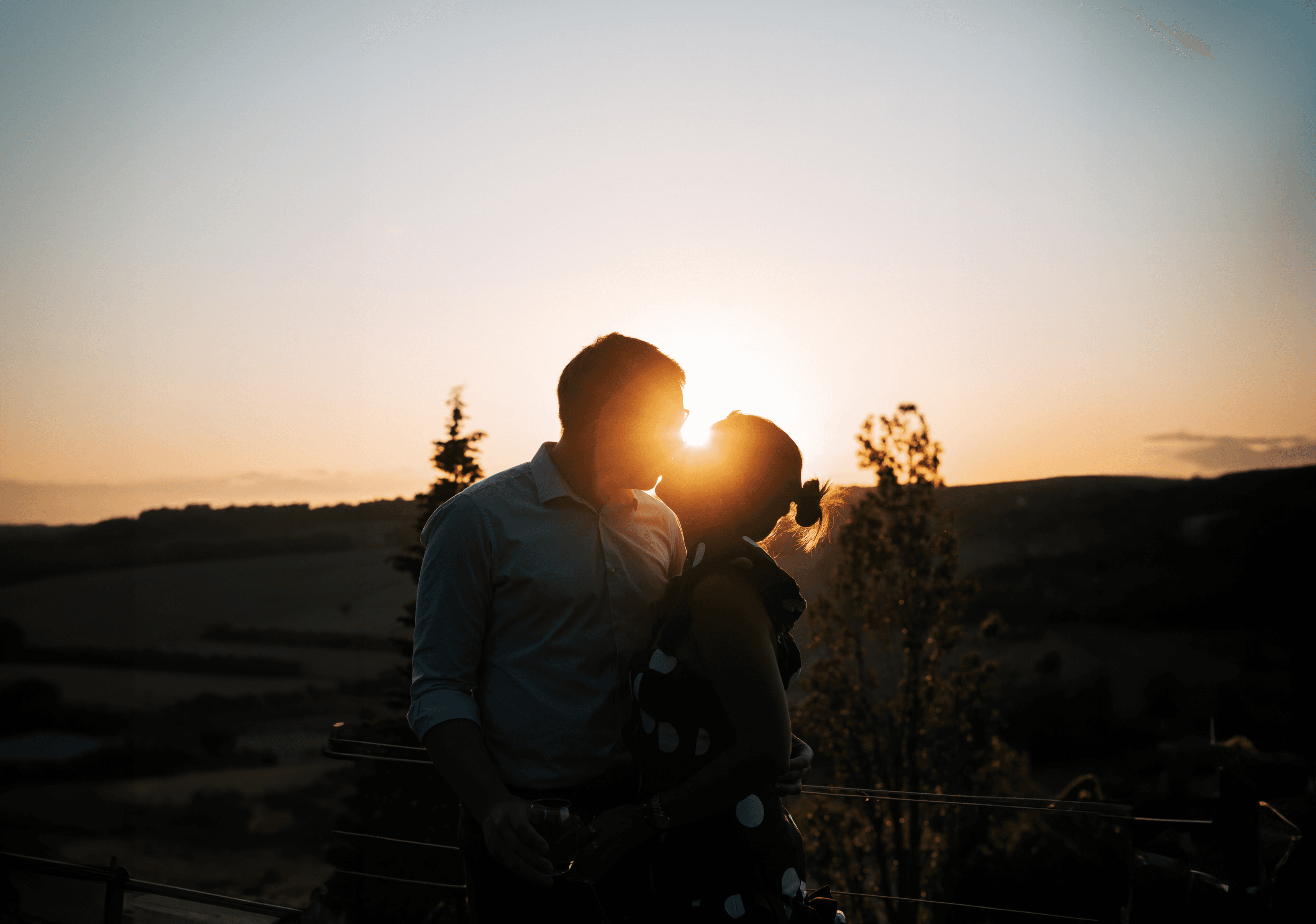 A bride and groom hold hands tenderly, the bride's bouquet of dried flowers hanging low. They stand in a softly lit, elegant interior, conveying romance.