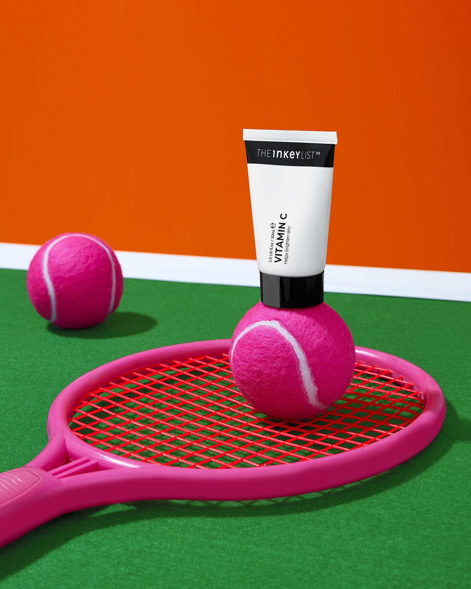 Vitamin C skincare tube on pink tennis ball and racket, orange background, green surface.