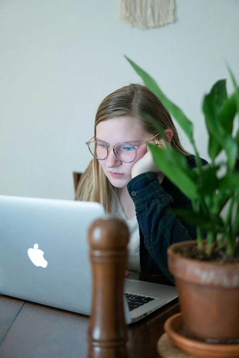 A woman with glasses studying at a laptop beside a houseplant.