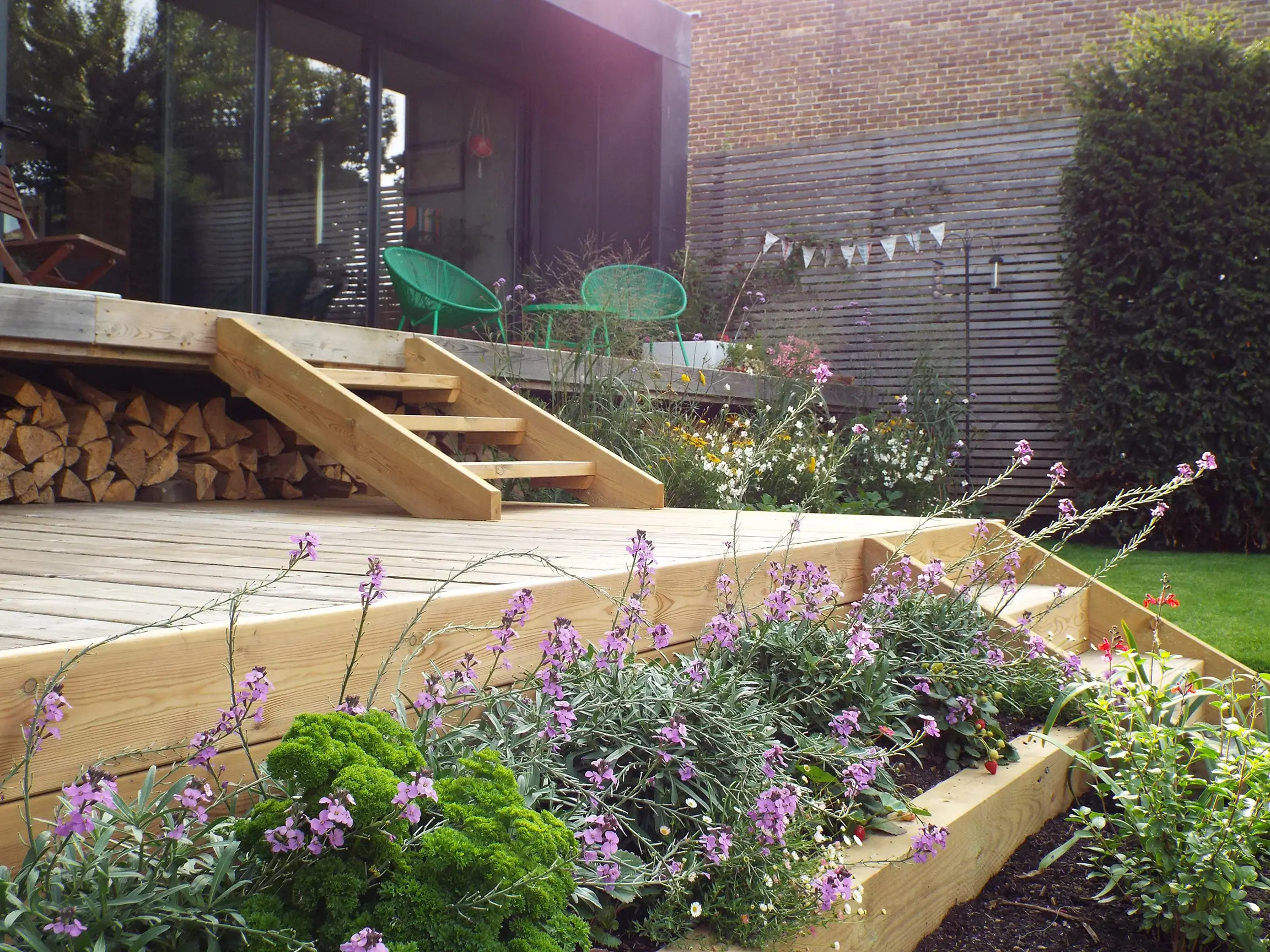 Sunlit garden scene featuring wooden steps leading up to a patio, surrounded by vibrant green plants and flowers.