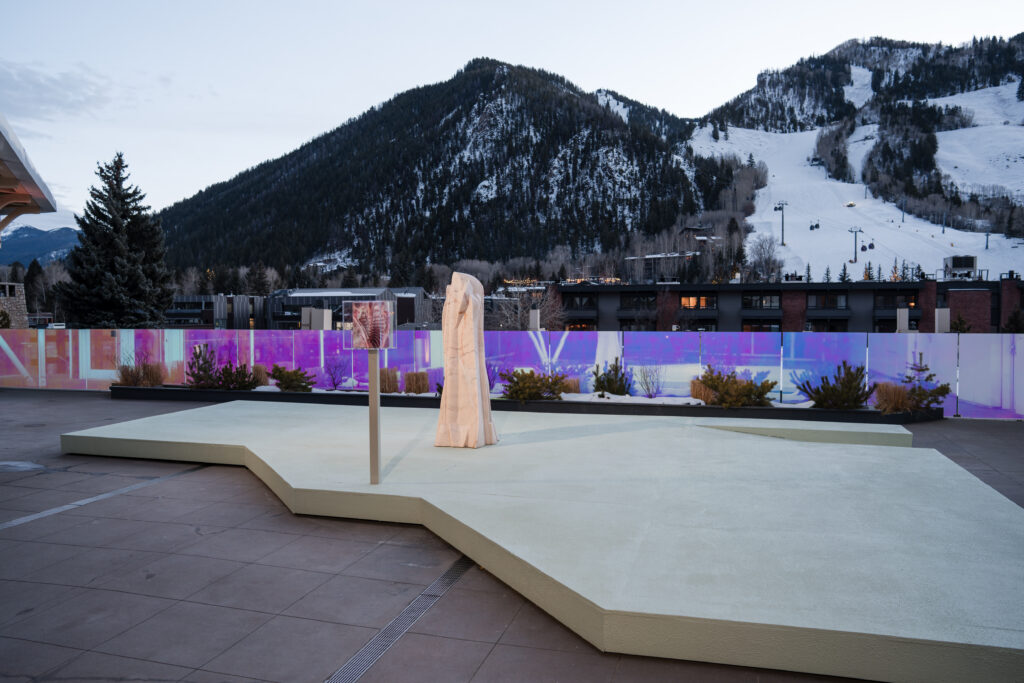 Outdoor installation on a pale platform beside an iridescent barrier, with snowy ski slopes behind it