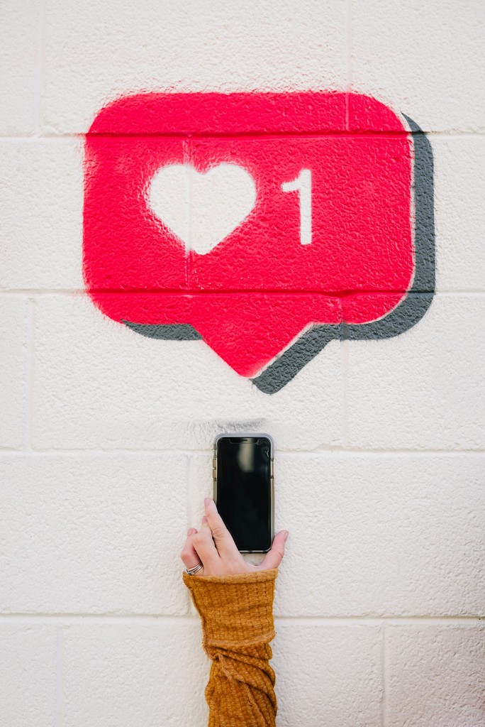 A hand holding a smartphone reaches up towards a painted social media "like" notification with a heart and the number 1 on a white brick wall, symbolizing the pursuit of online validation and engagement.