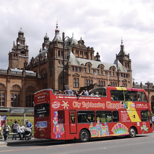 A red double-decker tour bus with "City Sightseeing Glasgow" parked in front of a historic, ornate building.