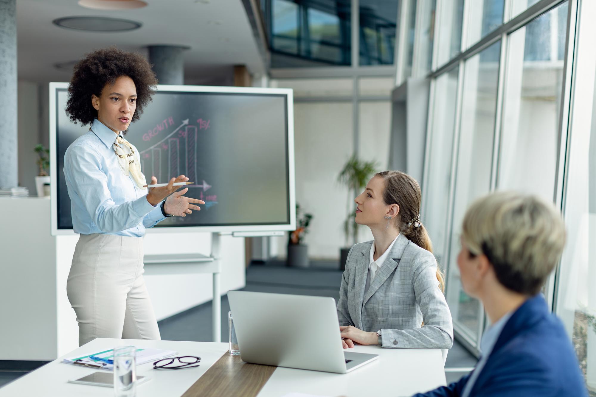 A woman stands in front of a meeting room display, showing a graph, talking to two other individuals about what's displayed while they listen attentively