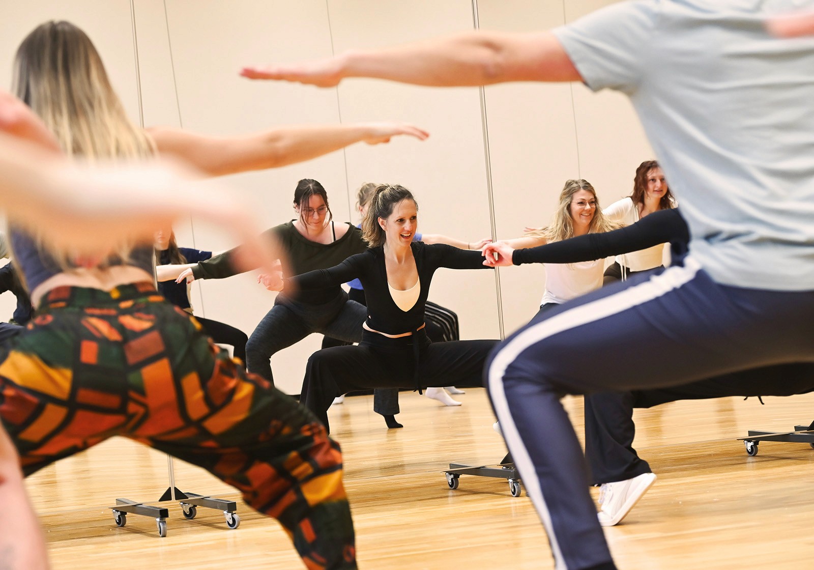 Eine Gruppe von Personen steht in der Abschlusspose eines Tanzes mit gestreckten Armen und Beinen in einem Trainingssaal.