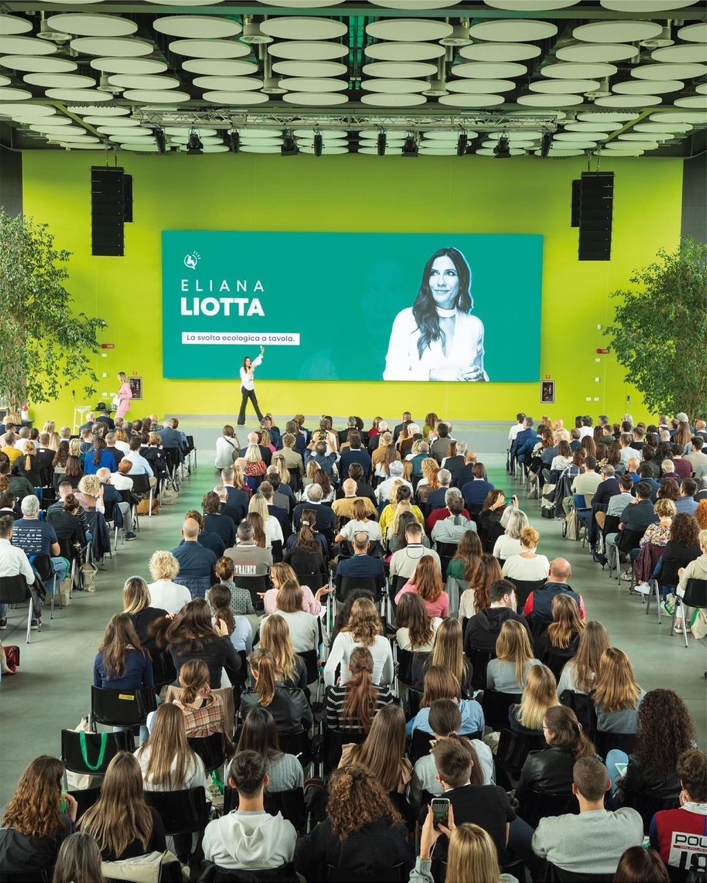 A young person with curly hair smiles at the camera while sitting in front of a laptop against a dark background.