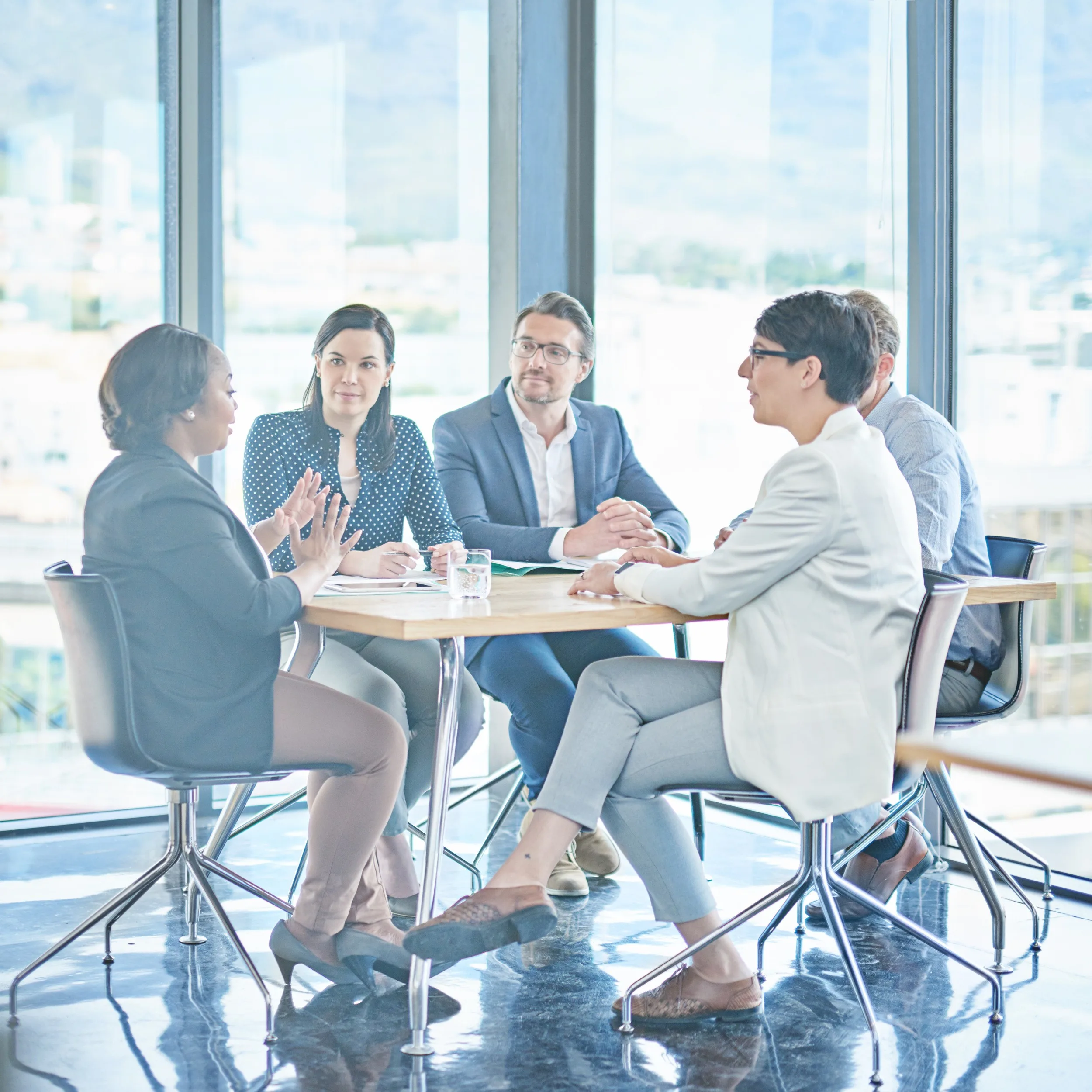 Five business professionals engage in a discussion around a wooden table in a high-rise office with expansive city views through floor-to-ceiling windows.