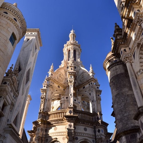 Un plafond voûté et orné avec des sculptures minutieuses, trois grandes fenêtres cintrées, et la lumière du soleil inondant une salle vide au sol en pierre.