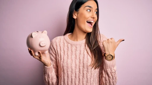 A woman in a light pink sweater is joyfully pointing and holding a small piggy bank against a pink background.