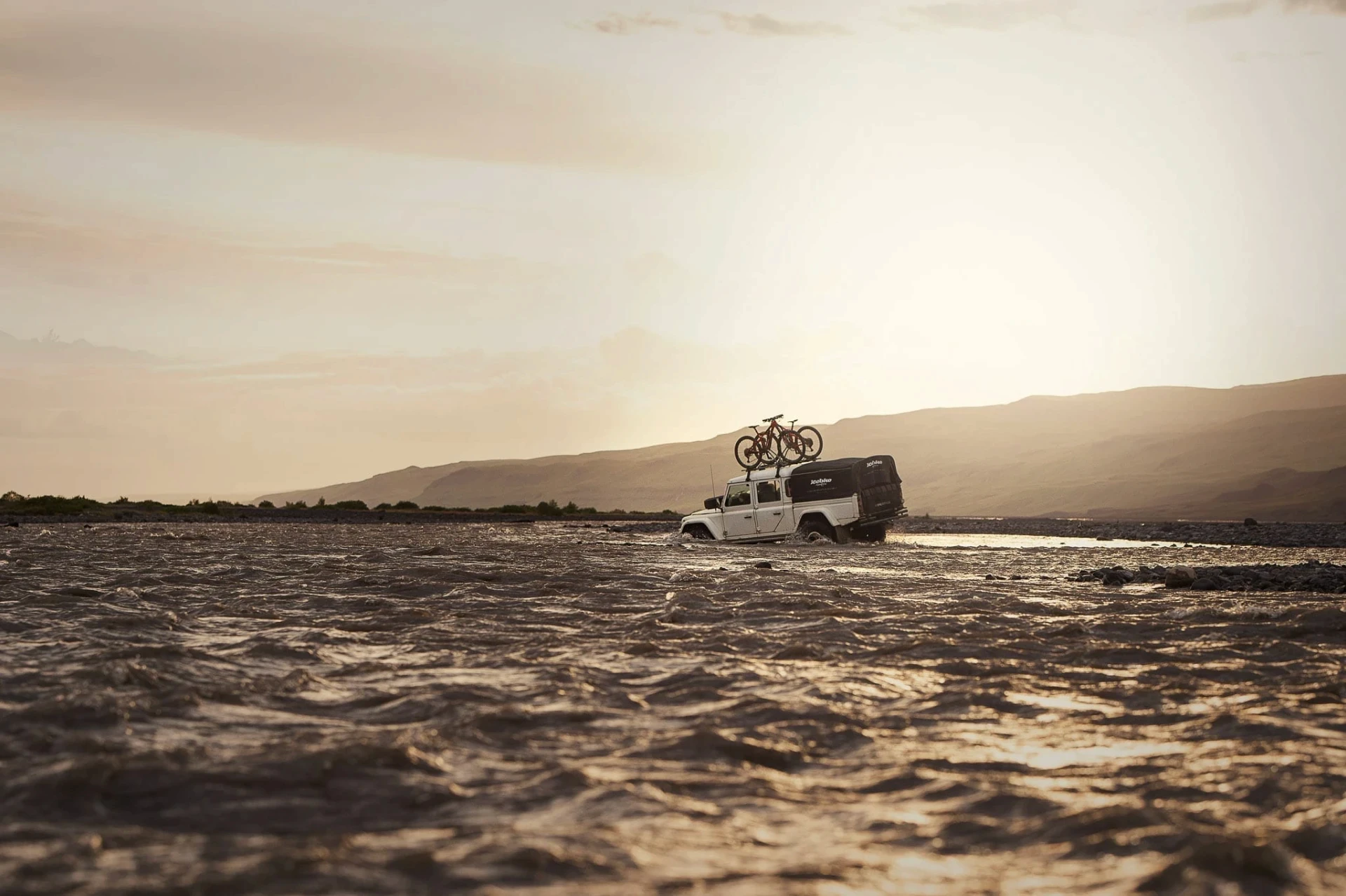 4x4 vehicle driving through a shallow river with bikes on the roof at sunset.