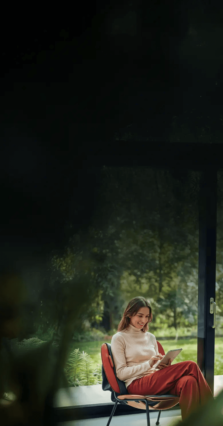 Check company name - Woman in turtleneck sweater and red pants smiles while looking at a tablet, sitting in a chair by a window.