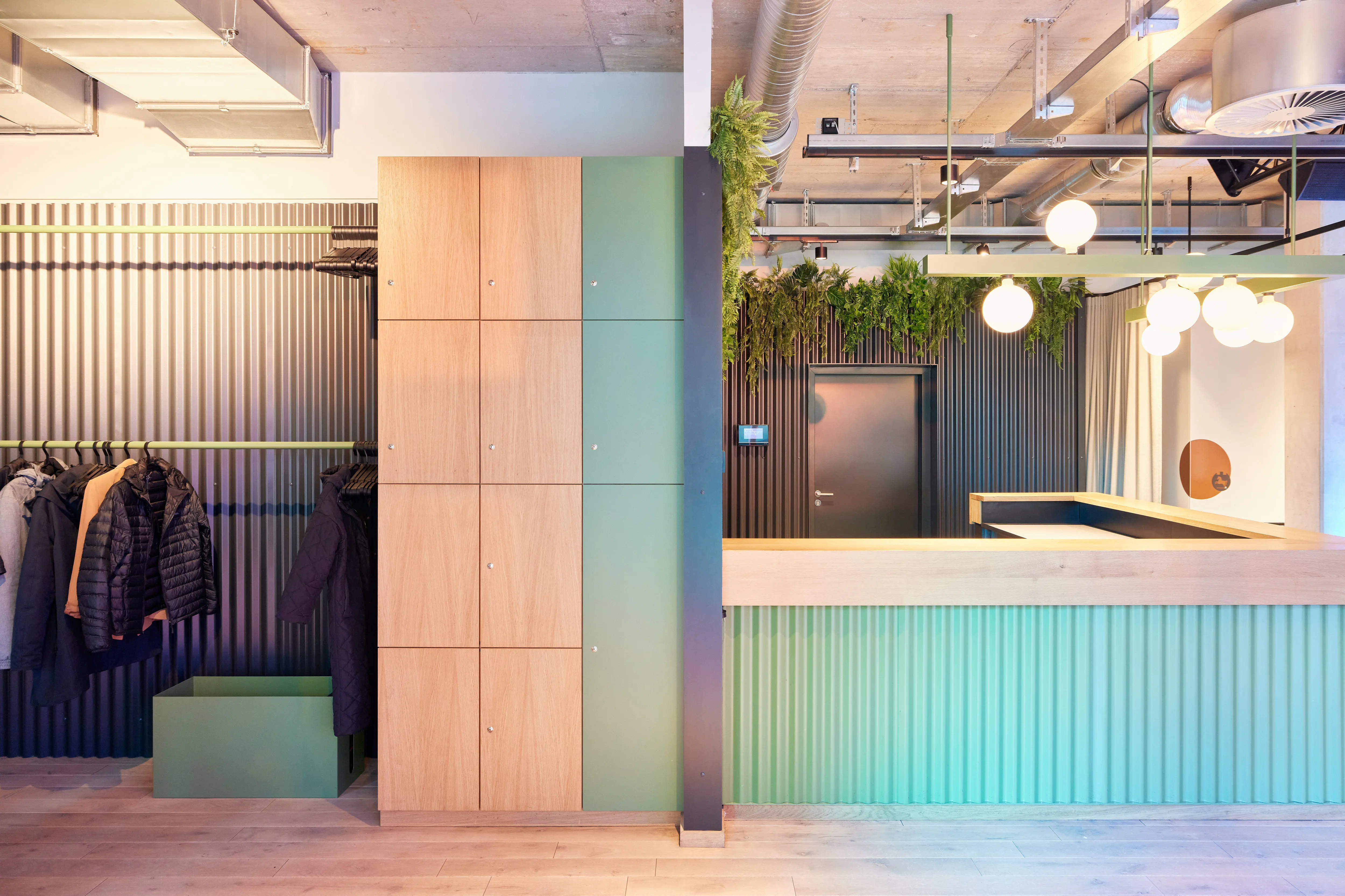 Entrance area of 381 EVENT SPACE with an open wardrobe in front of dark corrugated walls, wooden lockers with mint-green doors and the turquoise reception counter behind.