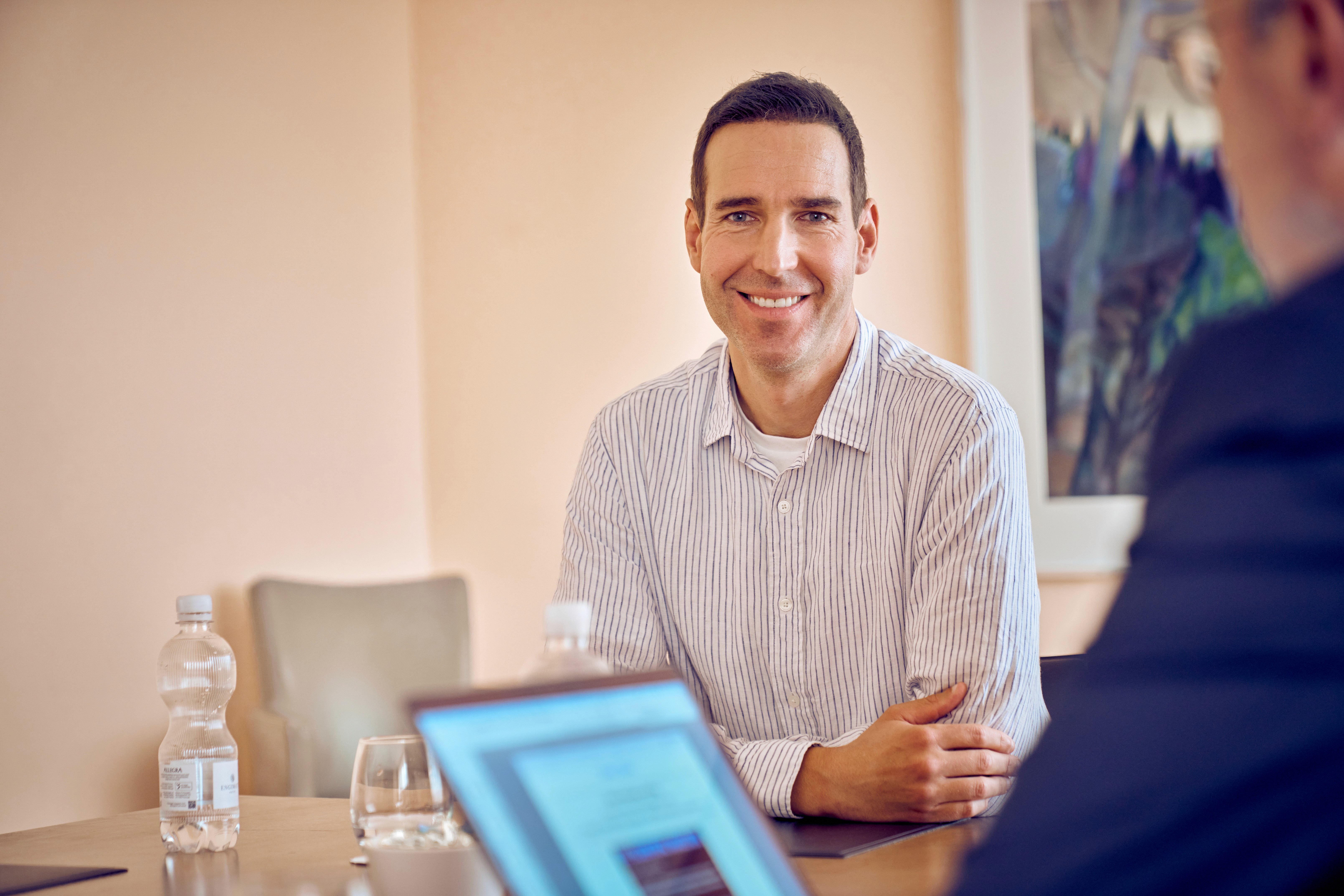 Zwei Büromitarbeitende sitzen an einem Holztisch, auf dem ein Laptop und viele Dokumente liegen. Die beiden Kolleg:innen geben sich einen High Five und lachen sich an.
