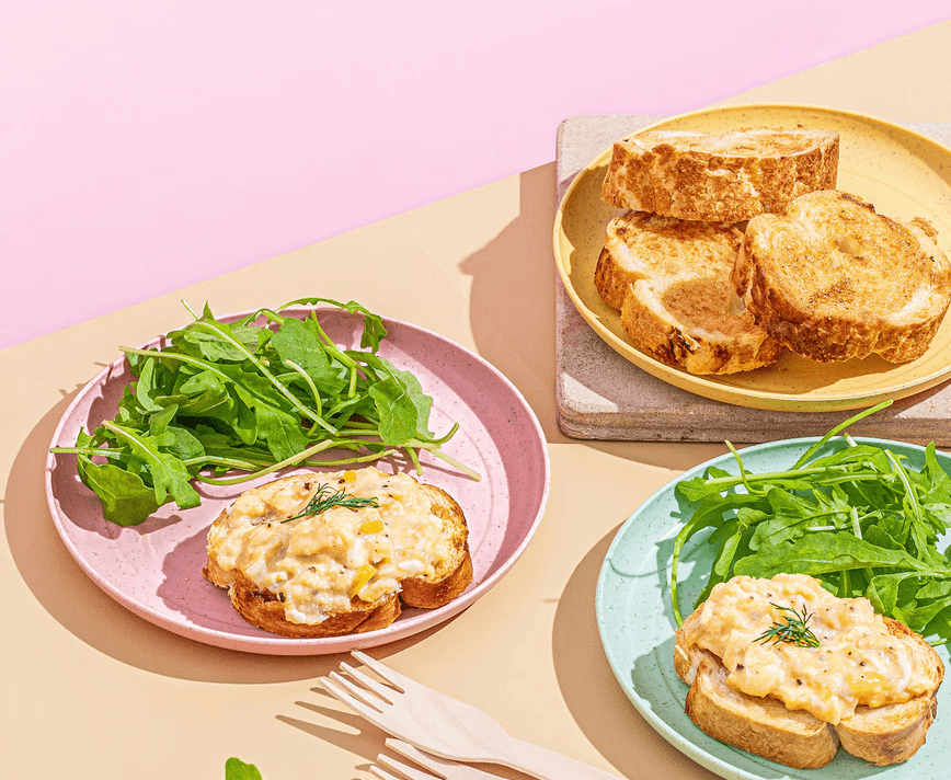 Creamy egg products on toasted bread with fresh arugula, served on pastel pink and mint green plates under bright, sunny.