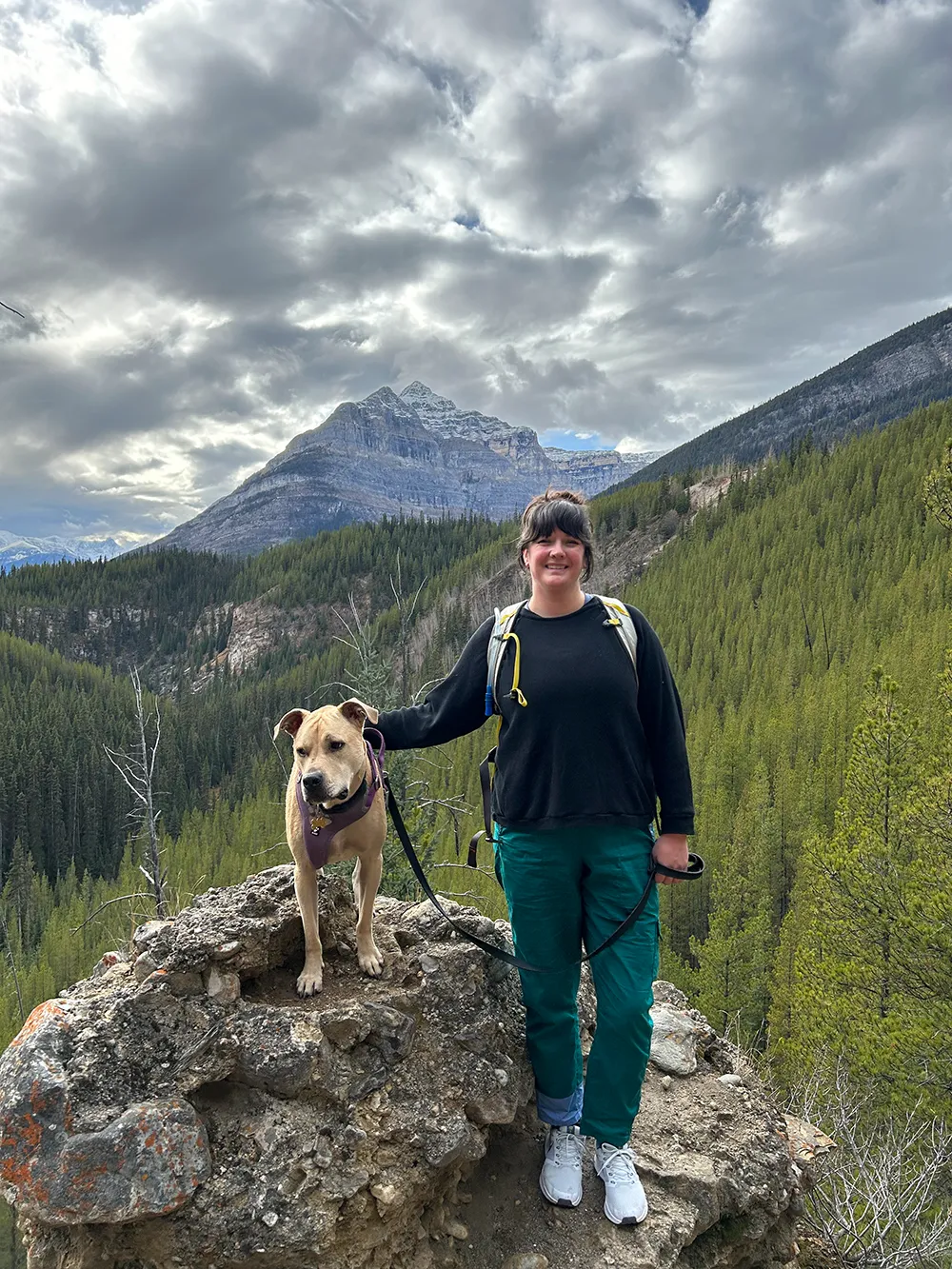 Psychologist standing in the mountains next to her dog with clouds in the distance