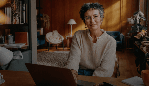 A woman with short hair and glasses sits confidently at a desk in a cozy, well-decorated room.