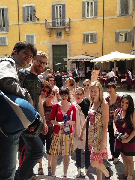 A photo of a group of people in the street while in the background people walk by a yellow building.