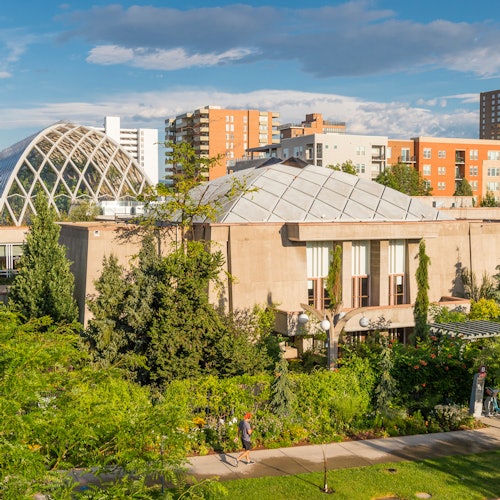 A building with a geometric dome roof, lush greenery in the foreground, and several modern buildings in the background.