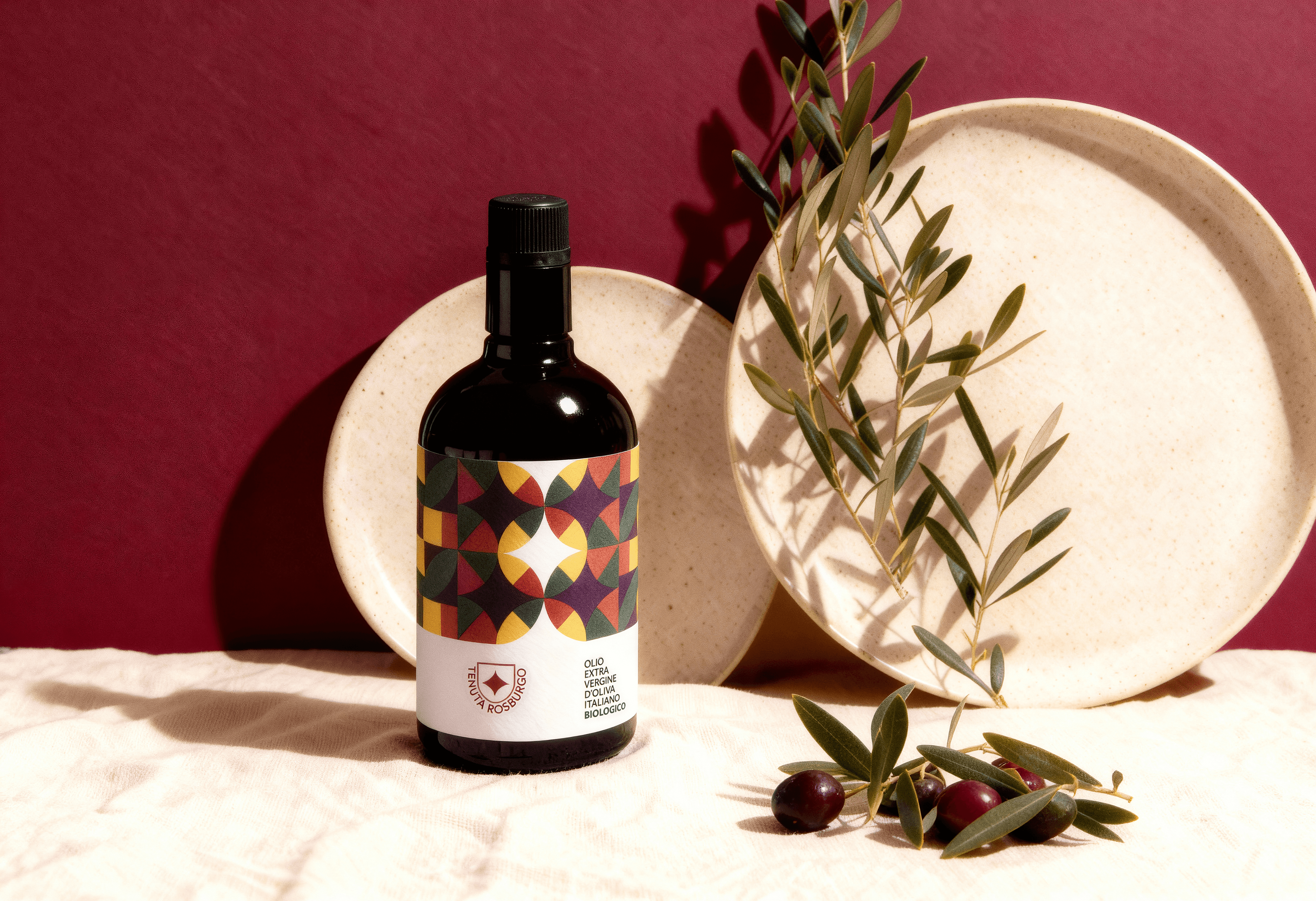 AI-generated product photography of Tenuta Rosburgo olive oil bottle with geometric label, plates, and olive branches on beige fabric. Created by Myrovia Labs for luxury food branding and AI packaging design.