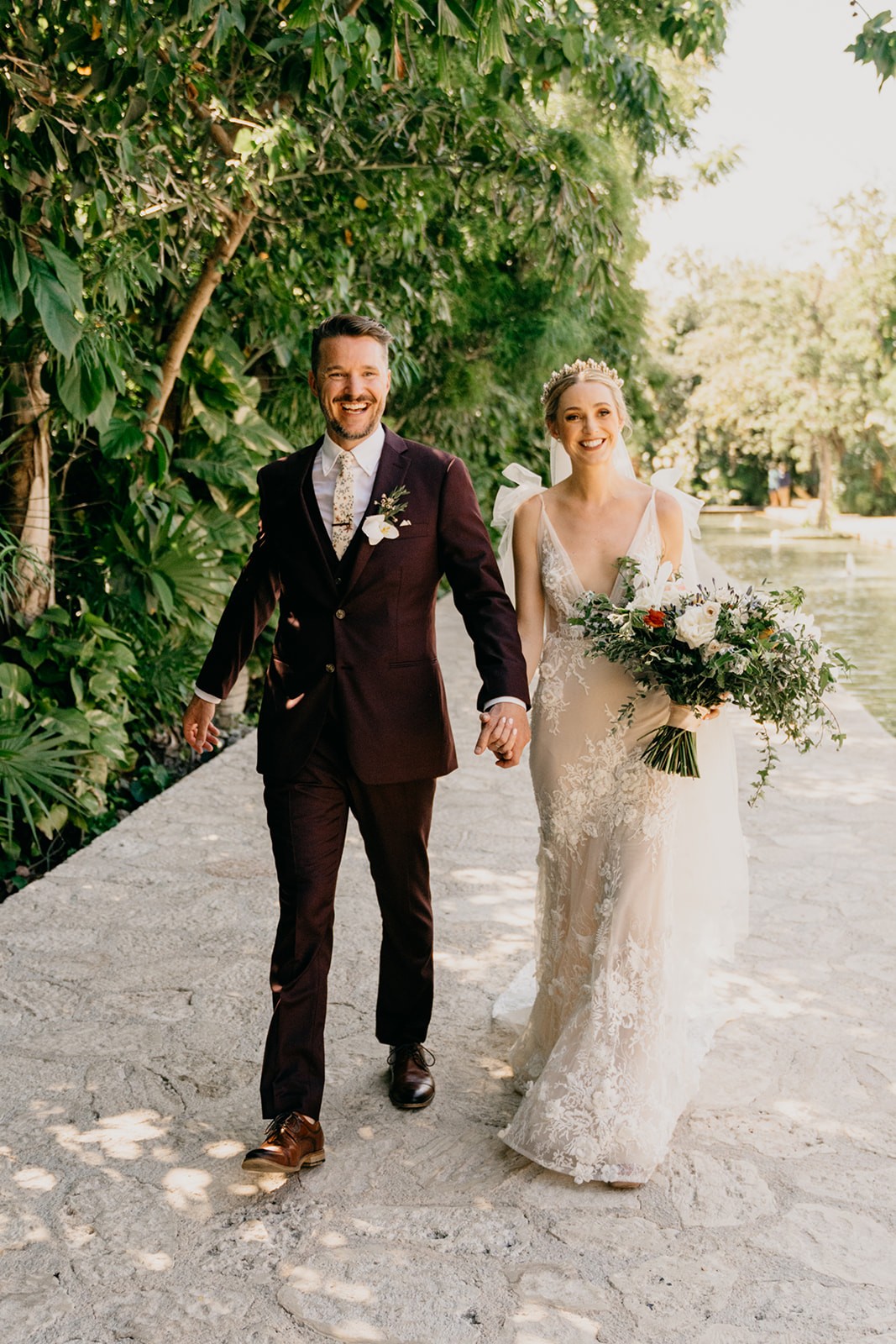 A bride and groom walking hand in hand outdoors, surrounded by greenery and a cheerful atmosphere.