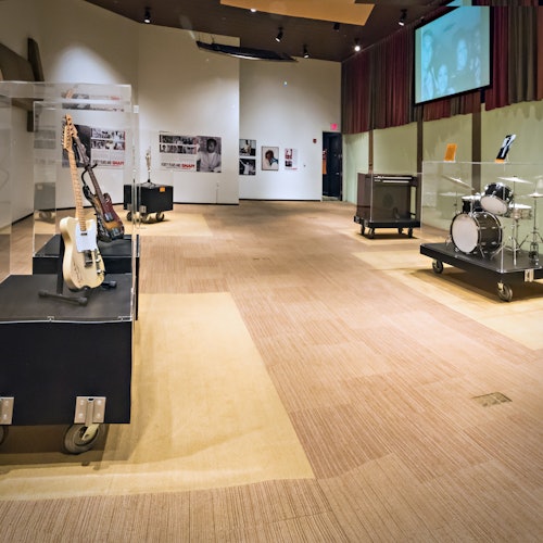 Muzejní expozice s elektrickými kytarami, bicí soupravou na pojízdných platformách a vintage hudebními plakáty na zdech pod měkkým osvětlením.