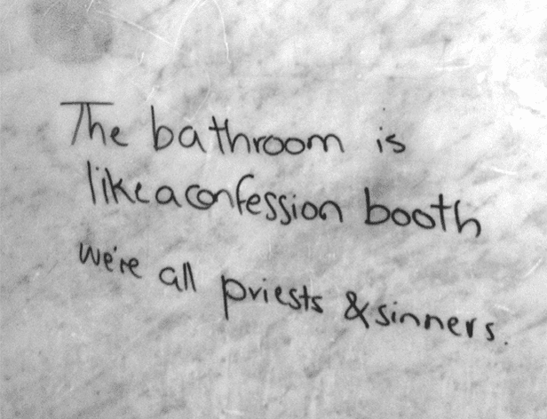The image shows a black and white photograph of a wall with handwritten text in black marker. The text reads, "The bathroom is like confession booth, we're all priests & sinners." The handwriting is informal and somewhat messy. The wall surface appears to have some texture or imperfections.