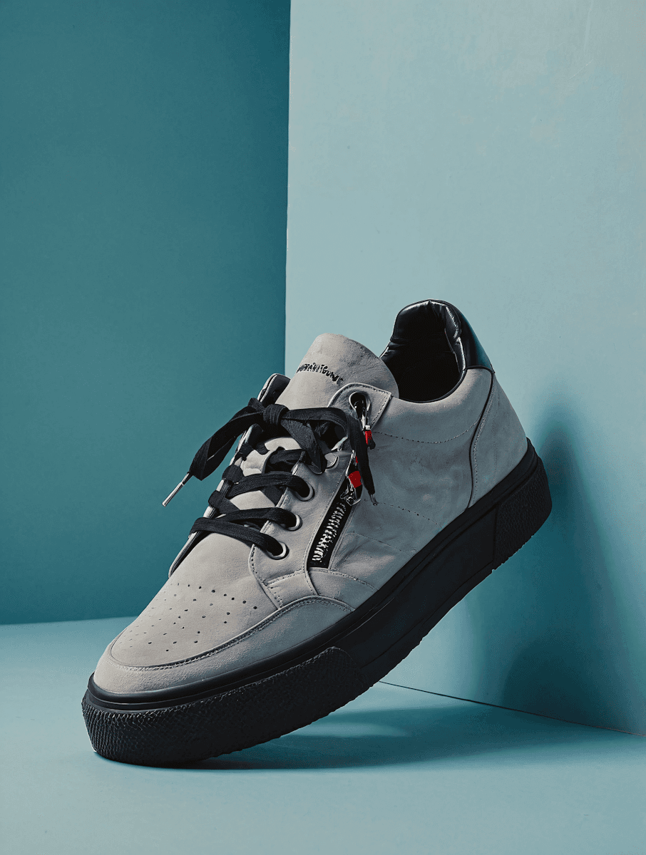 A grey suede low-top sneaker featuring a side zipper and black laces.