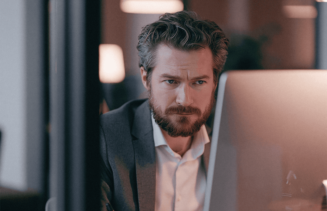 Homem de barba e paletó trabalhando concentrado em frente a uma tela de computador de mesa. Blog post Inspira.