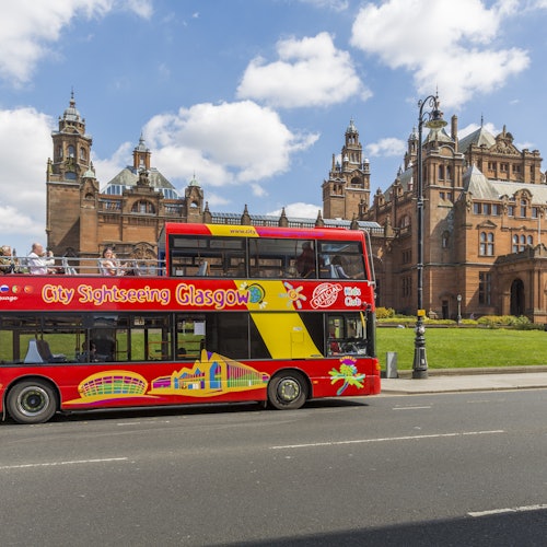 A red double-decker sightseeing bus with "City Sightseeing Glasgow" drives past a historic building under a partly cloudy sky.