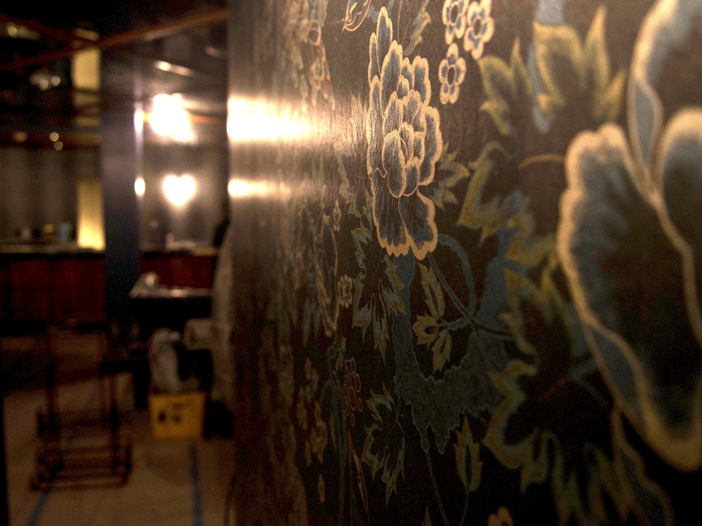 Close view of gold-toned floral wall covering with a softly lit wall sconce in the background.