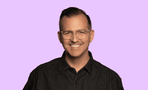 Headshot of a smiling man with glasses wearing a black button-up shirt against a light purple background.