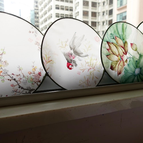五个装饰性的圆形扇子，饰有花卉和鸟类图案，陈列在窗台上，窗外是城市景观。