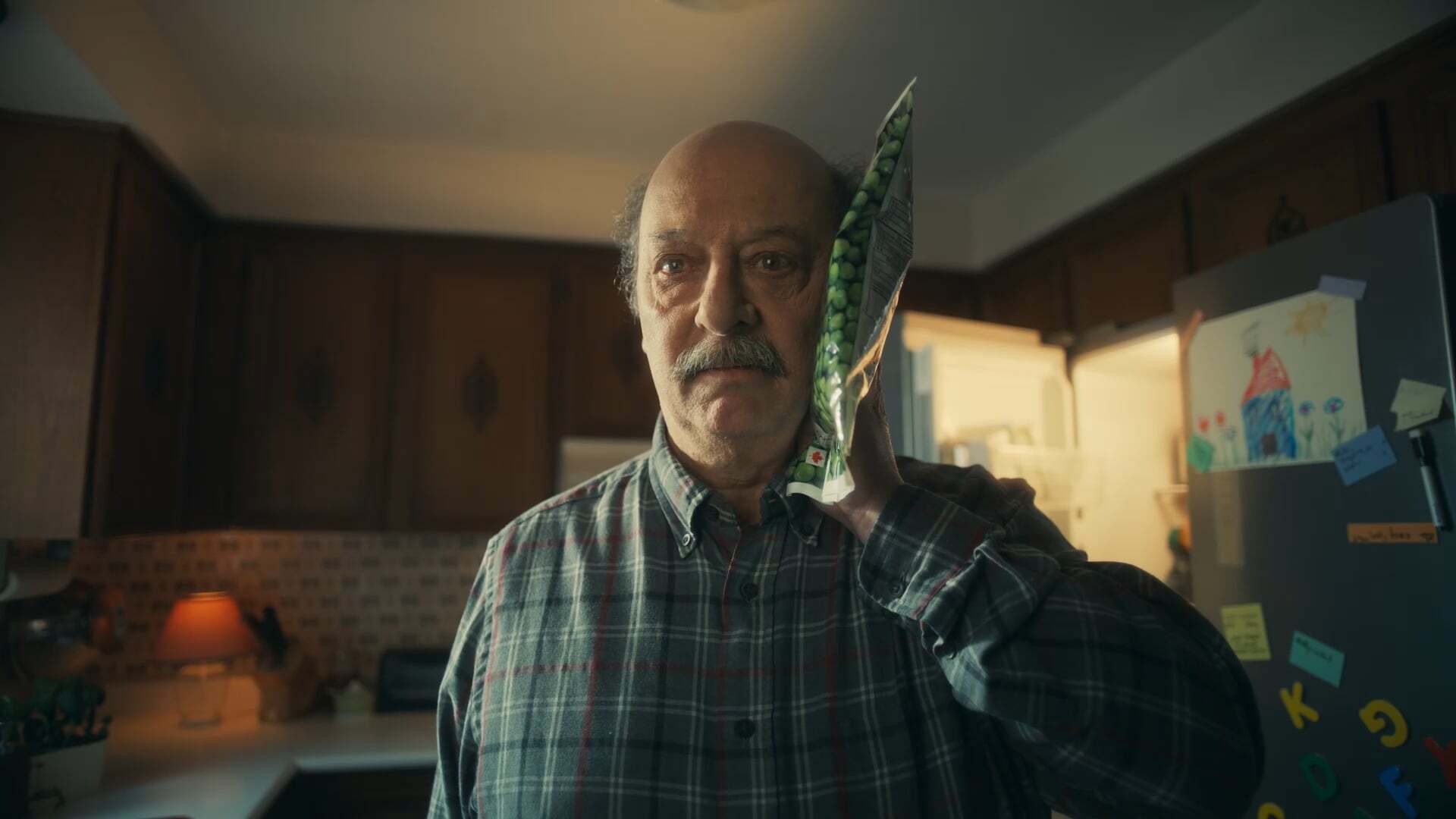 An older man holding a bag of frozen peas to his head in a dimly lit kitchen