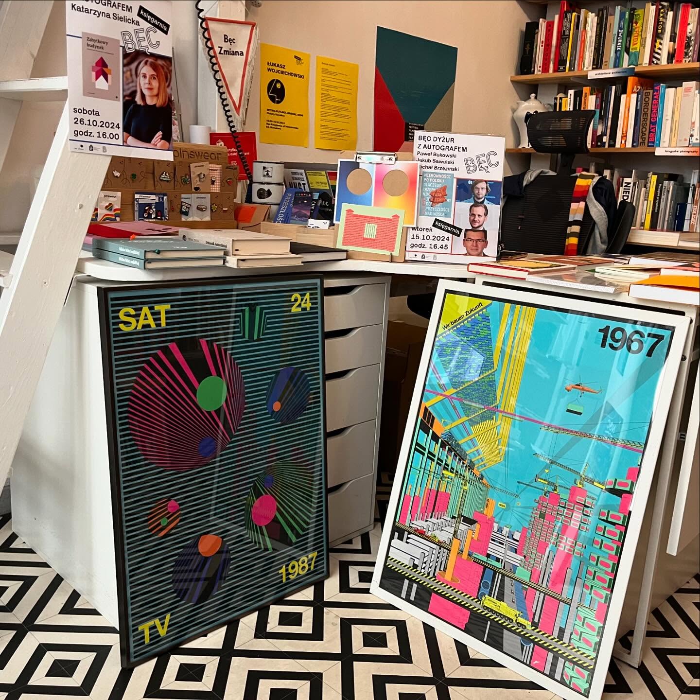 Colorful posters leaned on the desk, with bookshelves in the background and a patterned floor visible.